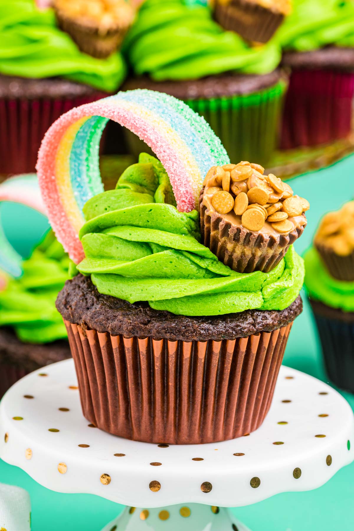 Chocolate cupcake with green frosting on a white cake stand. Decorated with rainbow and pot of gold candy..
