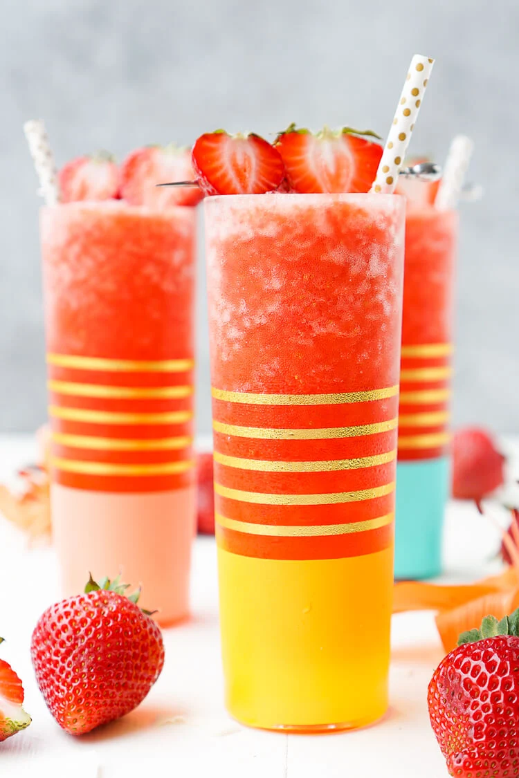 Photo of three gold striped glasses with frozen strawberry cocktail in them with gold polka dot paper straws garnished with fresh strawberries.