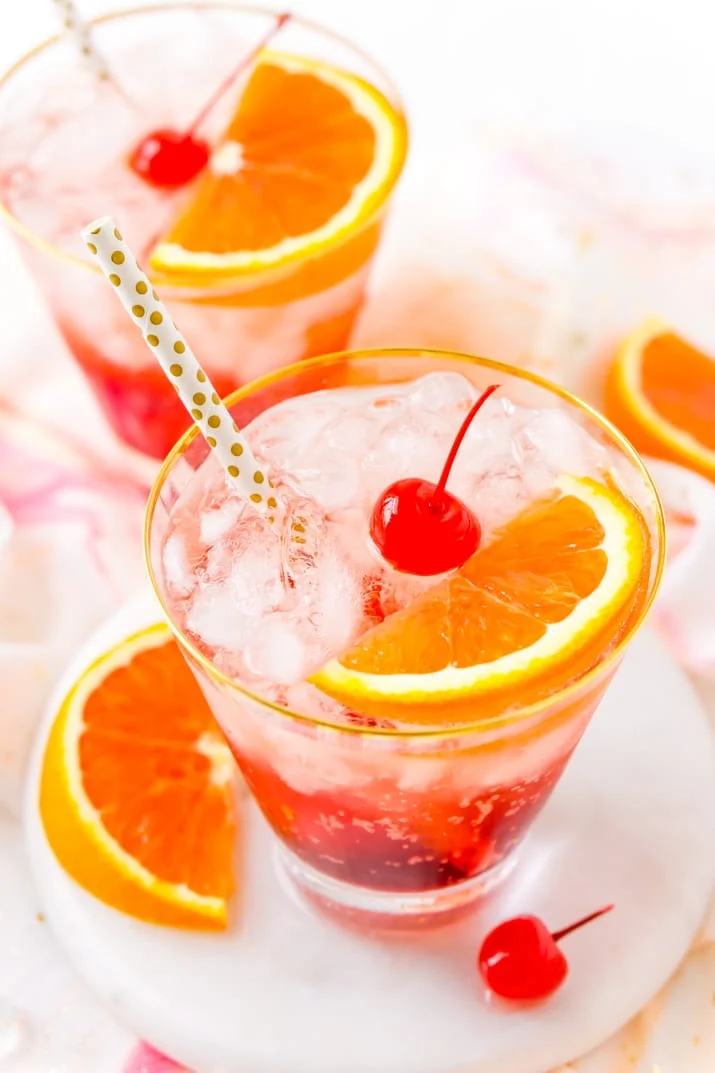 Two glasses filled with shirley temple drinks with gold polka dot straws and cherries and orange slices in them.