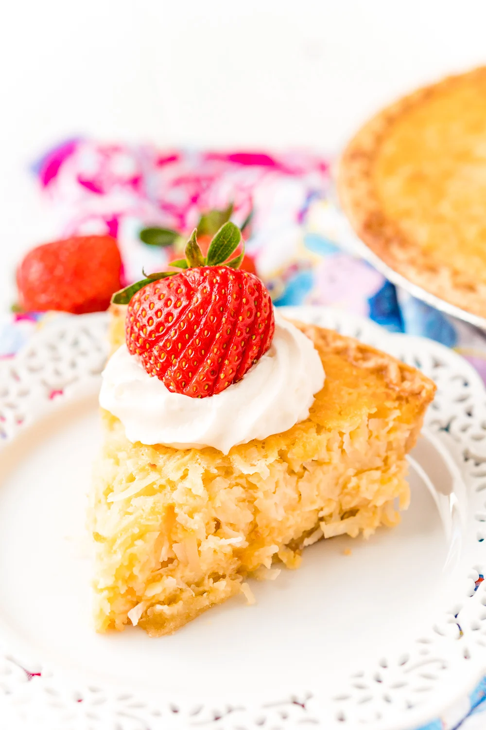 Slice of Coconut Custard Pie on a white plate with whipped cream and a strawberry on top.