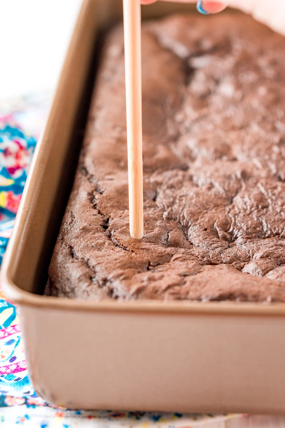 Chocolate cake in a 9x13-inch pan with a woman's hand holding a wooden skewer poking holes in the cake.