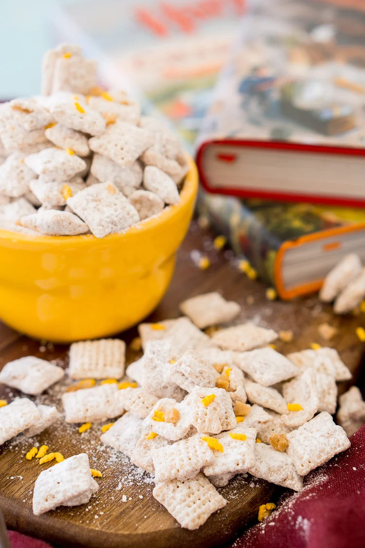 Butterbeer flavored muddy buddies scattered on a tables with a bowl behind them and Harry Potter books.