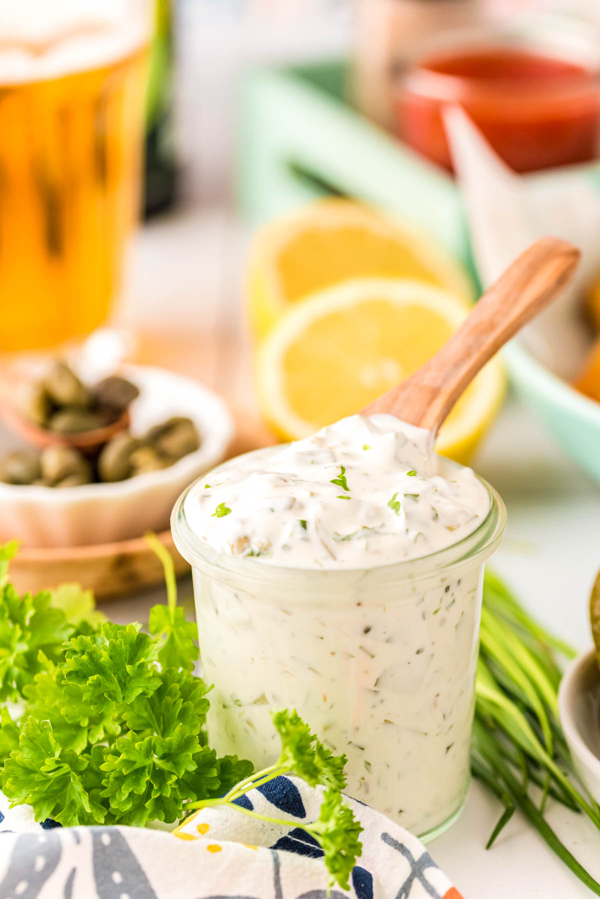 Tartar Sauce in a jar with a wooden spoon sticking out of it with ingredients in the background.