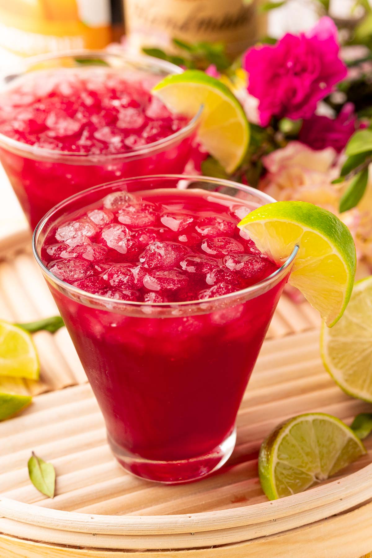Close up photo of two woo woo cocktails on a wooden serving tray with lime and flowers in the background.